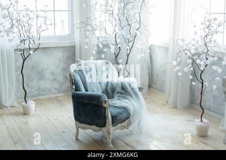 Elegant blue velvet armchair draped with sheer white fabric, surrounded by decorative white trees in a softly lit room. Creates luxurious and ethereal Stock Photo
