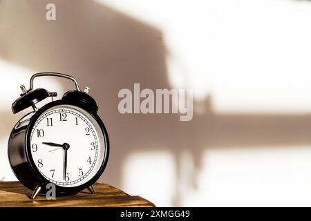 Retro-style black alarm clock on a wooden table with shadows on a white wall. Warm, vintage decor. Stock Photo