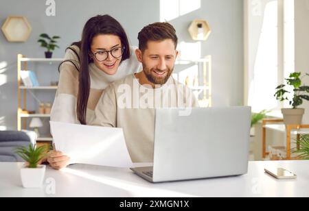 Cheerful young couple using laptop and analyzing their finances with paper document Stock Photo