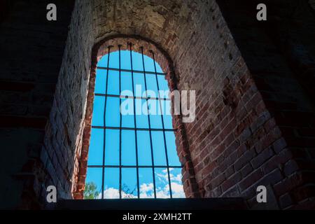 View from inside an old brick building showing an arched window with iron bars, revealing a blue sky with clouds. The rustic brick and iron bars creat Stock Photo