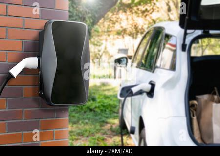 Charging electric car next to house with charging station on the wall. Stock Photo