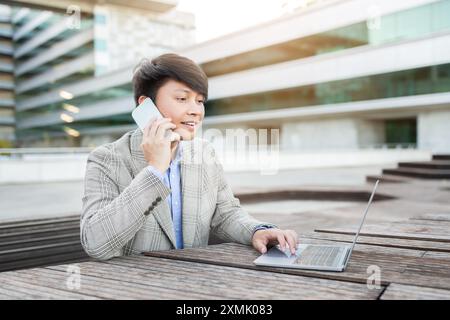 Young Man Talking on Phone, Using Laptop Outside Modern Building Stock Photo