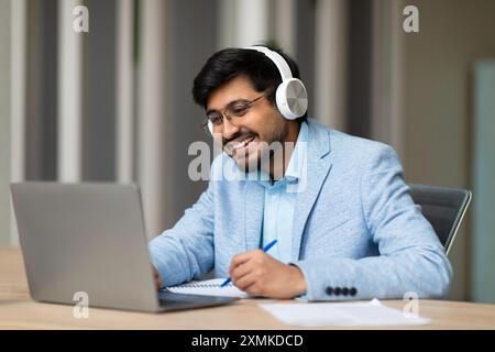Smiling Businessman Wearing Headphones While Using Laptop and Taking Notes Stock Photo