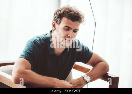 A thoughtful caucasian man with tousled hair sits on a chair in a bright, modern room. He wears a dark blue polo shirt and a silver wristwatch. The sc Stock Photo