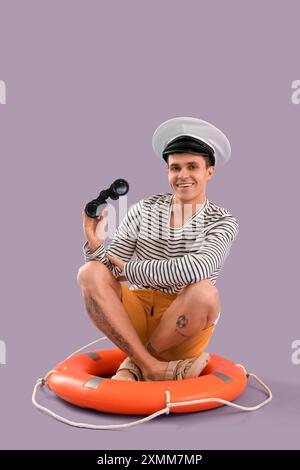 Young sailor with binoculars sitting on ring buoy against lilac background Stock Photo