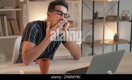 Sleepy student. Video lesson. Boring training. Tired young man at online lecture on laptop sitting at desk yawning in light living room home interior. Stock Photo