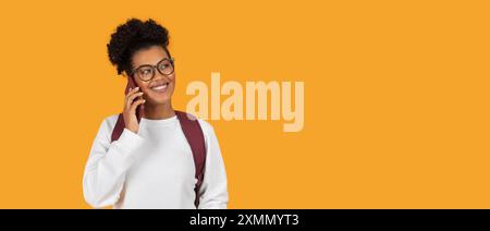 Smiling Young Woman Talking On Phone With Backpack Against Yellow Stock Photo