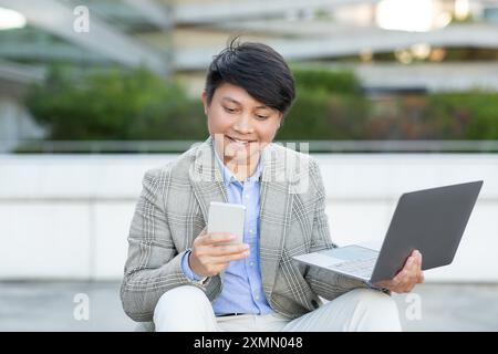 Smiling Man Using Phone and Laptop in Urban Setting Stock Photo