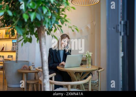 Businesswoman working at laptop in modern cafe, smiling talking on phone sits at table, multitasking Stock Photo