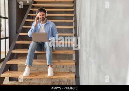 Man Working on Laptop While Wearing Headphones on Wooden Staircase Stock Photo
