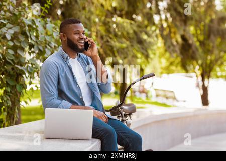 Handsome African Hipster Guy Talking On Cellphone And Using Laptop Outdoors Stock Photo