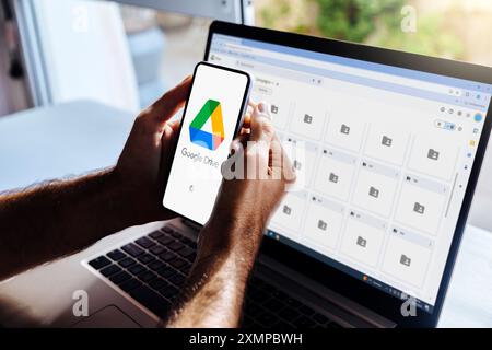 Google Drive on mobile phone and computer. Man holding a smartphone with cloud storage app on the screen. Laptop at background showing google folders Stock Photo