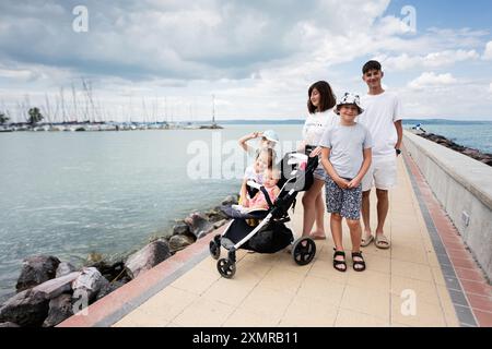 Happy family with children in stroller enjoying a leisure walk by a scenic marina with sailboats on a cloudy day. Stock Photo