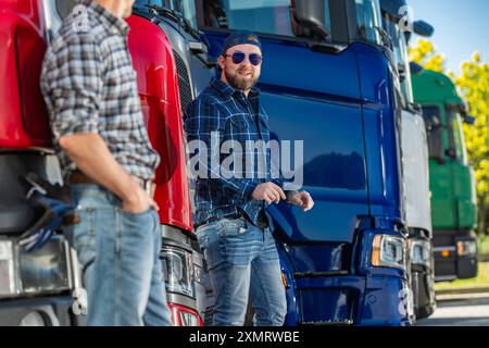 Two men in casual attire converse by a row of colorful trucks in a transportation yard. The bright sun casts a warm light as they engage in conversati Stock Photo