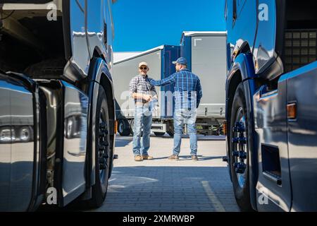 Two truck drivers engage in a conversation about logistics while standing between parked trucks in a transport yard. The bright blue sky adds to the v Stock Photo