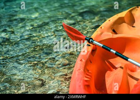 A vibrant orange kayak is partially submerged in crystal-clear water, showcasing colorful pebbles beneath. The lightweight paddle rests against the ka Stock Photo