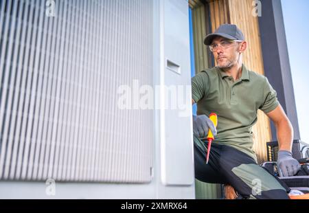 A technician in a green polo shirt and cap performs maintenance on an air conditioning unit. He uses a screwdriver and looks focused while working out Stock Photo