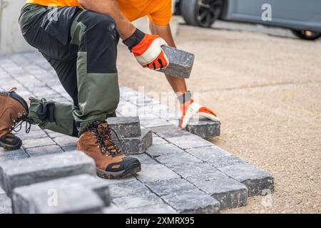 A construction worker is kneeling on the ground, carefully placing paving stones in a driveway. Wearing gloves and sturdy boots, they focus on arrangi Stock Photo