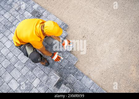 A construction worker is carefully placing gray paving stones onto a prepared surface, ensuring alignment and stability. Dressed in an orange shirt an Stock Photo