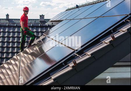 A technician in a red shirt and helmet stands on a slanted rooftop, carefully installing solar panels. The sun reflects off the shiny panels, illumina Stock Photo