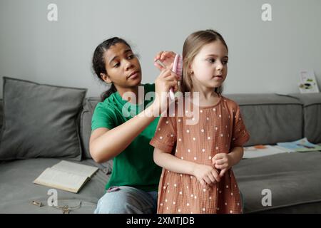 Cute teenage baby sitter brushing long thick hair of adorable child with plastic hairbrush while sitting on sofa behind little girl Stock Photo