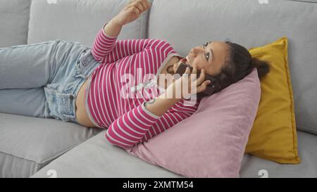 A young hispanic woman lies on a couch at home, talking on the phone, embodying relaxation and casual lifestyle. Stock Photo