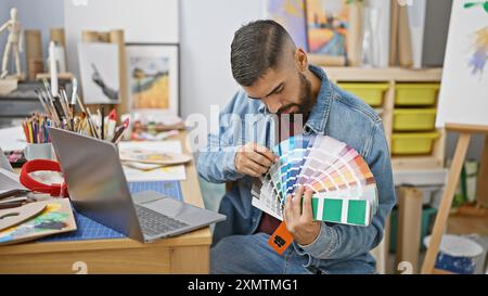 A bearded man examines color swatches in a creative studio with art supplies and a laptop nearby. Stock Photo