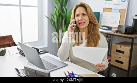 Confused hispanic woman answering phone call holding documents in modern office interior. Stock Photo