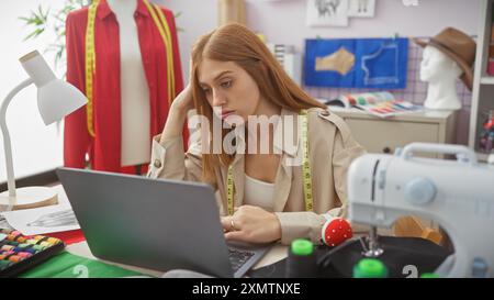 A focused woman tailor with measuring tape around neck works on laptop in a modern atelier surrounded by sewing items. Stock Photo