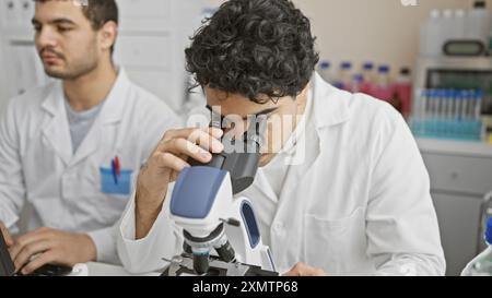 Two men in lab coats working in a laboratory, one using a microscope and the other taking notes. Stock Photo