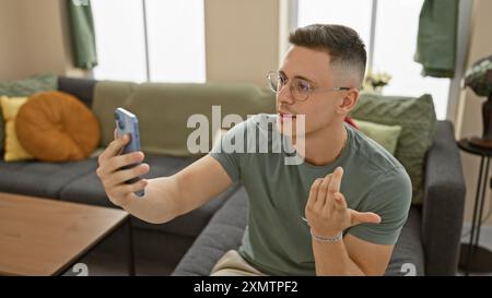 A young hispanic man in glasses takes a selfie indoors with a focused expression on his face, sitting on a couch. Stock Photo