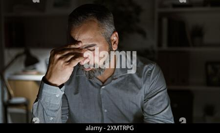 Stressed senior hispanic man with beard in office at night pondering over problems Stock Photo