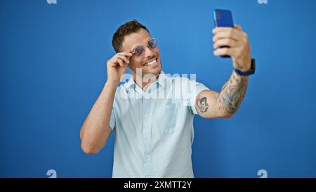 Handsome man with beard and tattoos smiles while taking a selfie, standing against a blue wall outdoors. Stock Photo