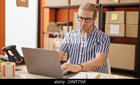 Handsome man with glasses working on a laptop in a warehouse office setting. Stock Photo