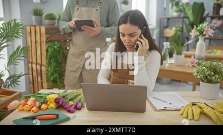 Woman talking on phone, man using tablet in flower shop with colorful tulips, plants, and work tools around. Stock Photo