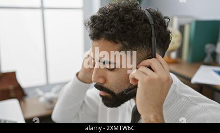 A pensive young hispanic man with a beard wearing a headset and working in a modern office. Stock Photo