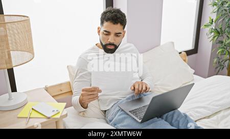 A young bearded man reads a document while sitting with a laptop in a bright cozy bedroom setting. Stock Photo