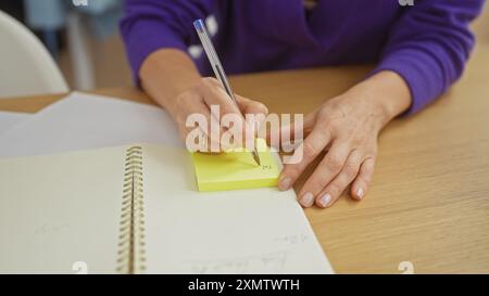 A woman writes on a sticky note at a wooden table indoors, illustrating planning or organization in a home setting. Stock Photo