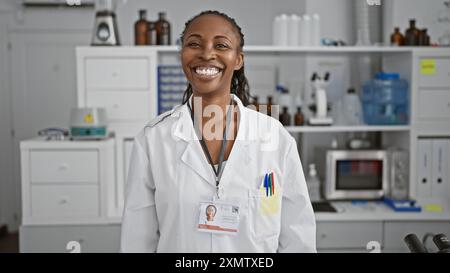 A smiling african american woman clinician with a badge stands confidently in a well-equipped laboratory room. Stock Photo