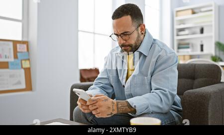 A bearded, young hispanic man in an office room using a smartphone while seated on a couch, looking engaged and stylish. Stock Photo
