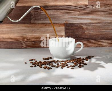 Coffee in white porcelain cup on white surface with wood panel background surrounded by coffee beans and shadows of maple leaves outside the scene Stock Photo