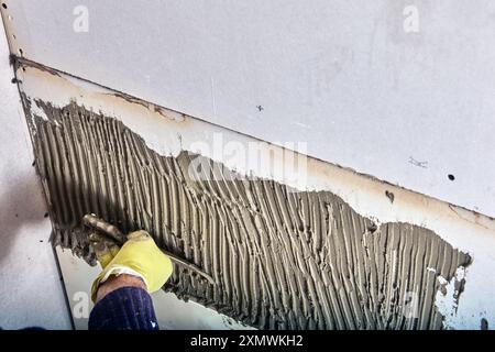 Construction worker tiler applies adhesive cement mortar to wall using notched trowel. Stock Photo
