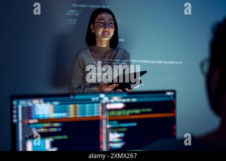 asian woman developers collaborating on a coding project, with one member presentation at code projected on the wall while the other works on multiple Stock Photo