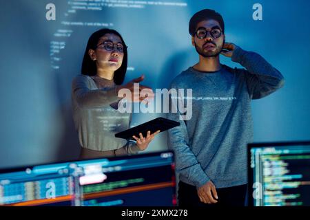 Asian Two software developers engaged in collaborative coding session, with code projected onto the wall and multiple computer screens displaying Stock Photo