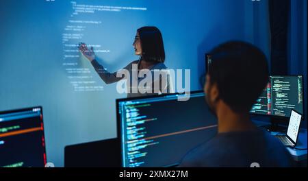 asian woman developers collaborating on a coding project, with one member pointing at code projected on the wall while the other works on multiple com Stock Photo