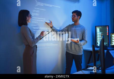 Asian Two software developers presentation in collaborative coding session, with code projected onto the wall and multiple computer screens displaying Stock Photo