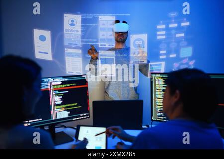 developer uses a VR headset to interact with projected interface designs, while colleagues discuss and code on screens at office Stock Photo