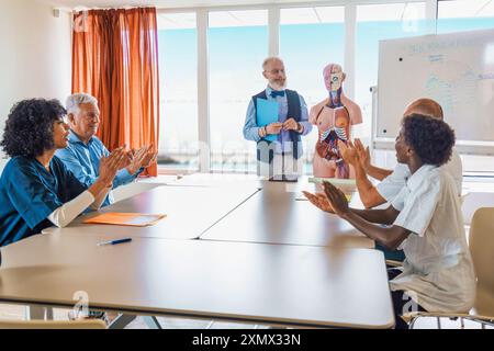 Senior professor teaching anatomy to diverse group of medical students using anatomical model. Multicultural class applauding during educational prese Stock Photo