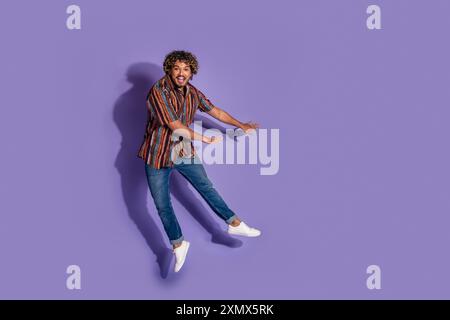 Full size photo of good mood impressed man dressed print shirt jeans jumping near offer empty space isolated on purple color background Stock Photo