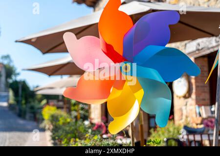 Close-up of a vibrant, multicolored pinwheel spinning in a sunny outdoor setting. The background features patio umbrellas and a charming garden, addin Stock Photo